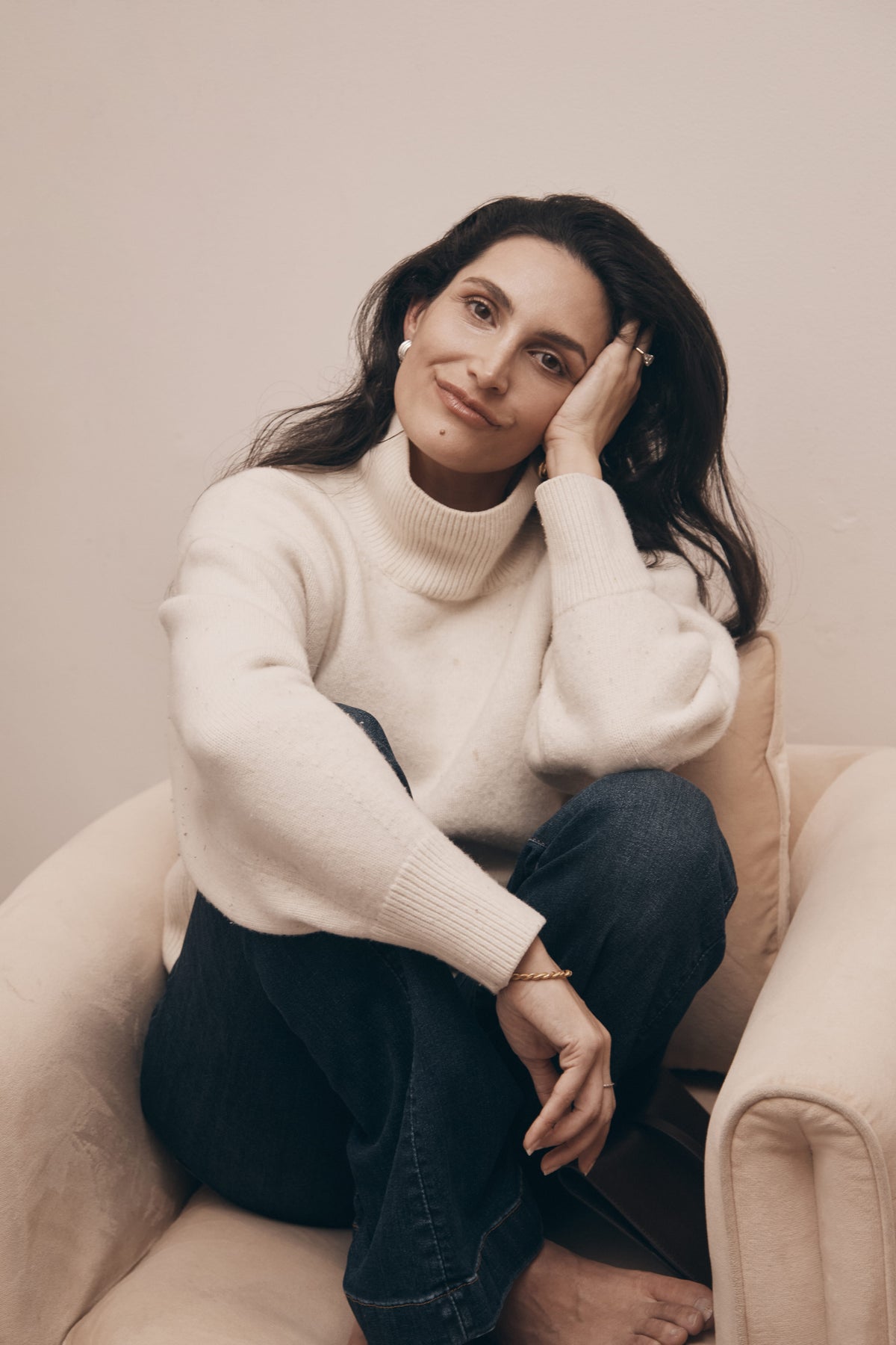 A woman with long dark hair sits barefoot on a beige armchair, wearing a cream-colored ribbed turtleneck sweater and dark blue jeans, resting her head on her hand against a neutral beige background.